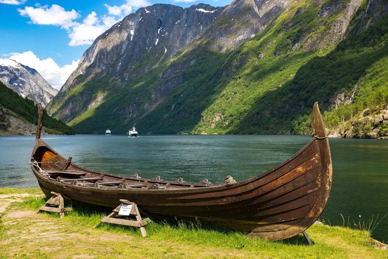 A wooden Viking ship rests on the grassy shore of a calm fjord, surrounded by steep green mountains and rocky cliffs.