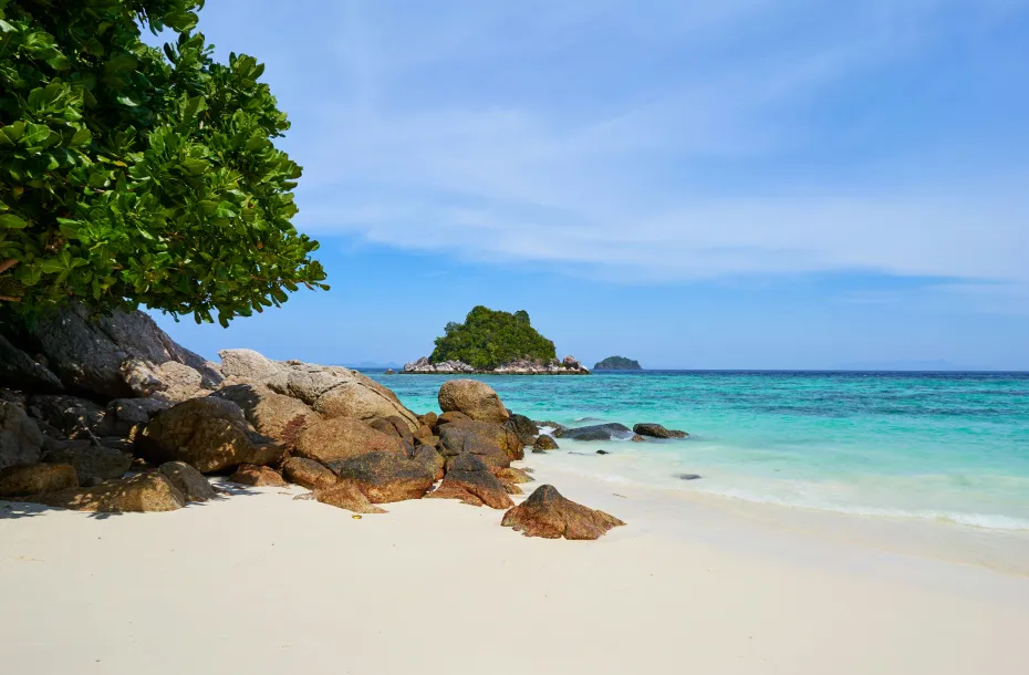 White sand beach in Koh Lipe, Thailand, with turquoise waters, smooth granite rocks in the foreground and a small green island in the distance.