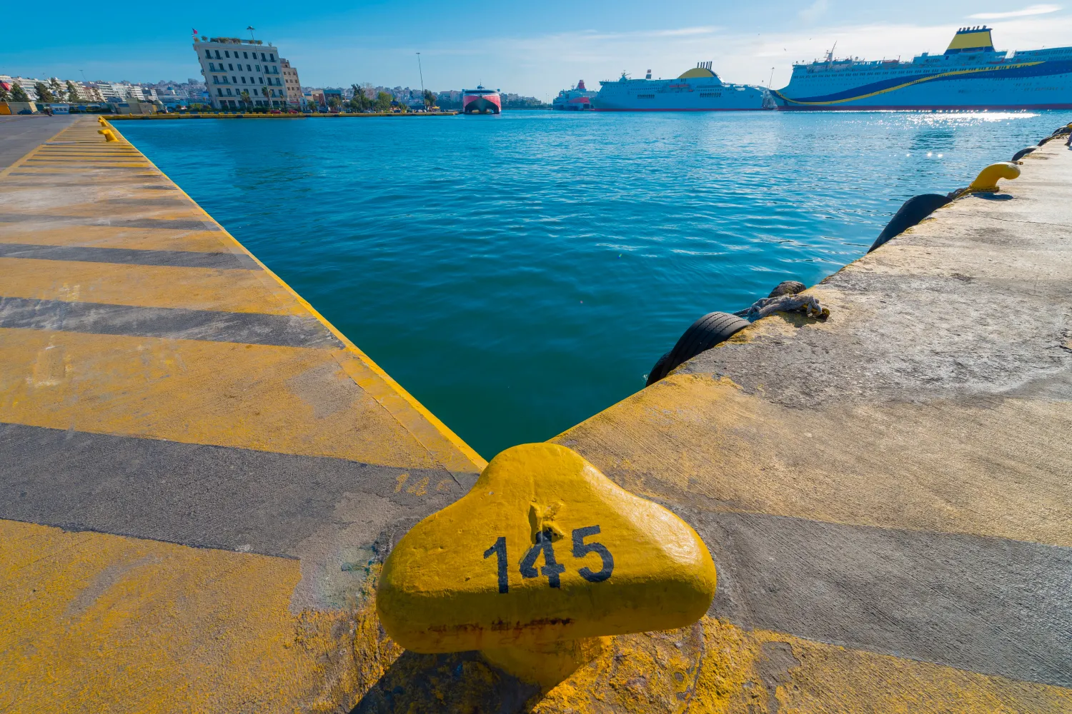 Piraeus port in Greece showing large passenger ferries docked at the pier with a yellow mooring bollard numbered 145 in the foreground under a clear sky.